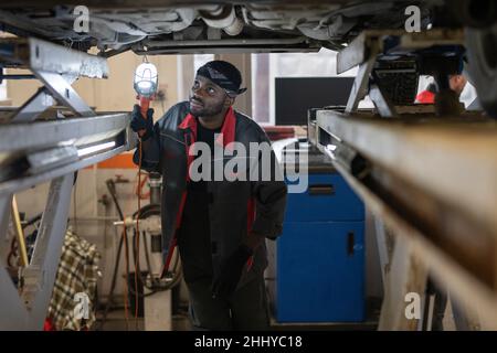 Jeune homme africain en vêtements de travail et gants tenant la lampe près de la partie inférieure de la voiture tout en se tenant sous le véhicule et en la vérifiant Banque D'Images