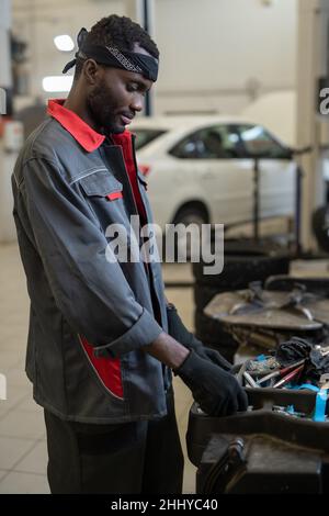 Un jeune réparateur ou technicien africain ganté en vêtements de travail prend l'outil à main nécessaire pour réparer une voiture cassée Banque D'Images