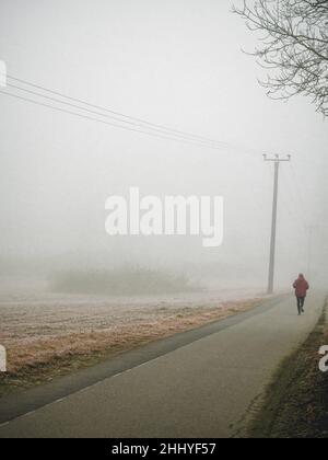 Un jogging solitaire court sur une route brumeuse.La route et les champs sont recouverts d'une épaisse brume le matin froid de l'hiver. Banque D'Images