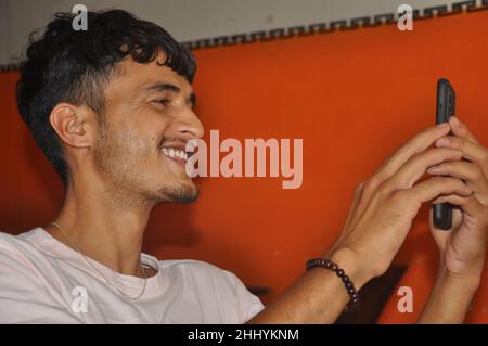 Vue rapprochée d'un jeune homme souriant tout en utilisant son téléphone pendant qu'il est assis à l'intérieur Banque D'Images