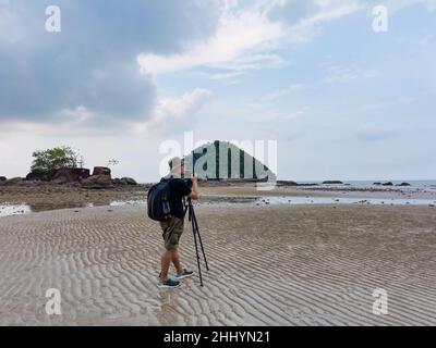 Photographe de la nature en action.Seul le voyageur tire le paysage marin avec un petit fond d'île.Backpacker prendre des photos sur la plage de sable à marée basse.FR Banque D'Images