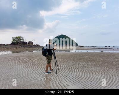 Photographe de la nature en action.Seul le voyageur tire le paysage marin avec un petit fond d'île.Backpacker prendre des photos sur la plage de sable à marée basse.FR Banque D'Images