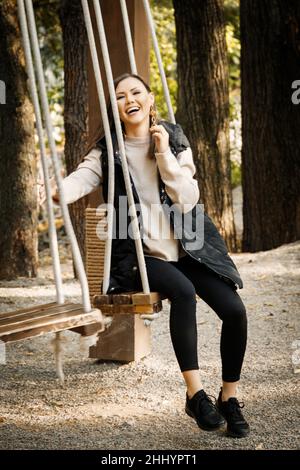 Jeune femme mongol heureuse avec cheveux bruns droits et maquillage, en faisant un swing parmi l'automne en plein air dans des vêtements chauds dans les couleurs de base dans un parc à l'extérieur Banque D'Images