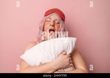 Belle jeune jolie femme mignon porter un masque de sommeil tenant un oreiller doux, naines comme veut dormir, triste de se réveiller tôt le matin isolé sur le b rose Banque D'Images