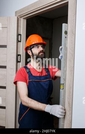 un travailleur avec une perceuse dans un casque orange installe une porte dans la maison Banque D'Images