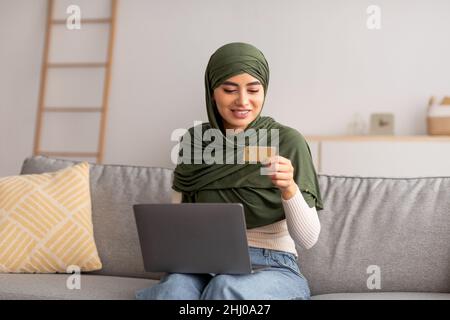Bonne femme arabe millénaire dans hijab en utilisant la carte de crédit et ordinateur portable, les achats en ligne sur le canapé à la maison Banque D'Images