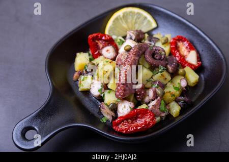 Salade chaude avec poulpe, pommes de terre, tomates séchées, câpres et citron dans une petite casserole noire. Gros plan, arrière-plan noir. Banque D'Images