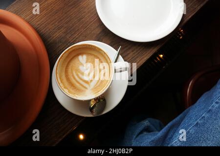 Service simple de cappuccino avec art floral en forme de cœur sur une table en bois sombre.Jean bleu d'une fille assise dans un café Banque D'Images