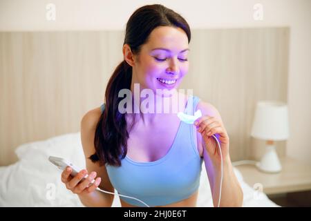 Une jeune femme utilise un complexe de blanchiment pour le blanchiment des dents à la maison.Lampe UV USB pour le blanchiment des dents. Banque D'Images