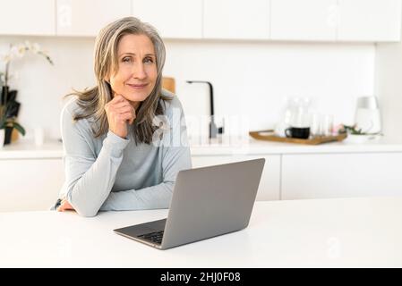 Gaie et attrayante, la femme mûre est assise dans la cuisine avec un ordinateur portable, une femme senior et sympathique qui discute en ligne, appréciant le travail à distance Banque D'Images