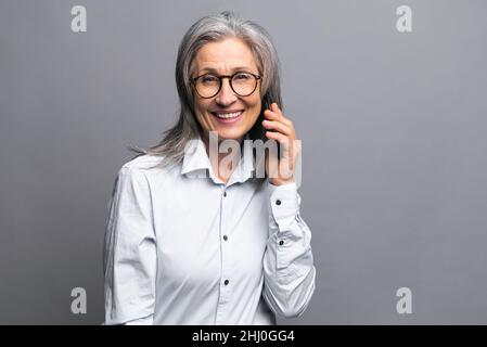 Femme d'affaires souriante et positive parlant au téléphone intelligent au-dessus du mur gris.Une femme de vente âgée a une conversation téléphonique.Une femme mûre parle sur son téléphone portable Banque D'Images