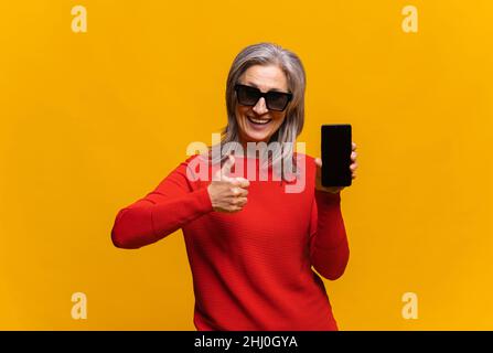 Photo horizontale d'une femme aîée hilarante en chandail rouge tenant un smartphone avec écran vide isolé sur jaune.Femme d'âge moyen à cheveux gris montrant le pouce vers le haut, comme un panneau Banque D'Images