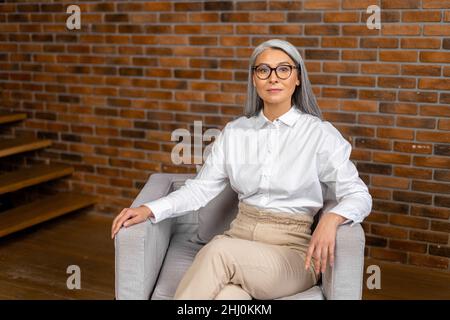 Portrait d'une femme sénior, leader, coach, femme d'affaires aînée en lunettes, assise au bureau et regardant avec assurance l'appareil photo Banque D'Images