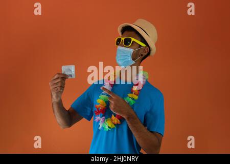 Homme noir en costume de carnaval et masque pandémique tenant un condom isolé sur fond orange.L'homme africain dans diverses poses et expressions. Banque D'Images