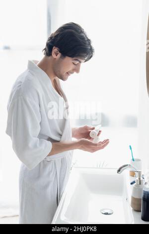 vue latérale de happy man en peignoir appliquant de la crème pour les mains près des articles de toilette sur le lavabo Banque D'Images