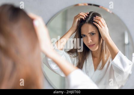 Bouleverser jeune femme debout près de Mirror et regardant ses racines de cheveux Banque D'Images