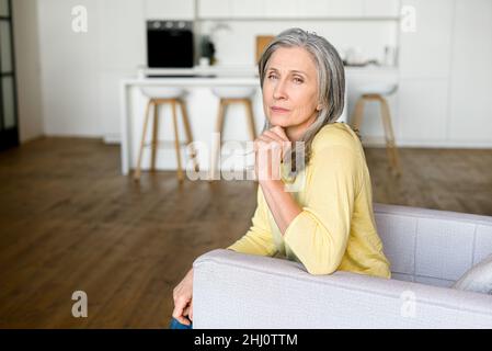 Femme sénior réfléchie assise sur le fauteuil seul et regarde la caméra avec une expression sérieuse du visage, menton reposant avec ponder de la main mature Banque D'Images