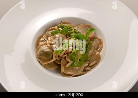 Raviolis ronds (tortelli) de farine complète avec pignons de pin et pesto génois et feuilles de basilic dans un plat blanc, gros plan Banque D'Images