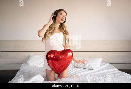 Saint-Valentin, Fête des femmes, anniversaire. Une jeune femme dans un casque est assise au lit et fête le 14 février avec un ballon en aluminium rouge en forme de coeur Banque D'Images
