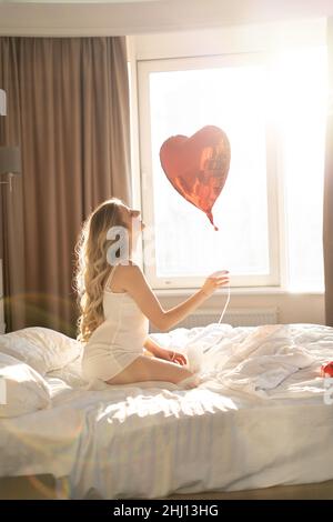 Belle jeune fille à la maison sur le lit le matin au soleil. Profitez de la fête de la Saint Valentin avec des ballons en forme de coeur rouge. Banque D'Images