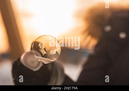 une fille tient une bulle de savon avec de beaux motifs de gel dans les rayons du soleil en hiver Banque D'Images