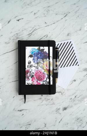 Bloc-notes noir au centre de la table en marbre, à côté d'une carte lumineuse à fleurs, d'une enveloppe blanche et d'un stylo élégant Banque D'Images