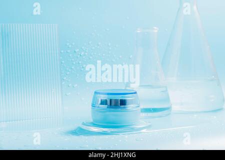 Crème cosmétique dans un pot, éclaboussures et gouttes d'eau sur fond bleu avec flacons en verre Banque D'Images