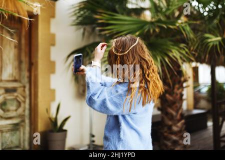 Femme méconnaissable en chandail bleu avec des cheveux de brunette en streaming avec le smartphone en main, le tournage de blog de médias sociaux dans un café extérieur moderne.Hipster Influencer en utilisant les réseaux sociaux. Banque D'Images