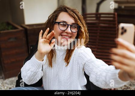 Bonne jeune caucasienne, millénaire ou la femme de génération z avec de longs cheveux de brunette streaming avec le smartphone en main, le tournage de blog de médias sociaux dans le café extérieur moderne.Hipster Influencer en utilisant les réseaux sociaux. Banque D'Images