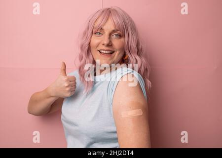 La femme vaccinée par coronavirus montre un signe de pouce avec bandage après avoir reçu le vaccin Covid 19 isolé sur fond de paroi rose. Banque D'Images