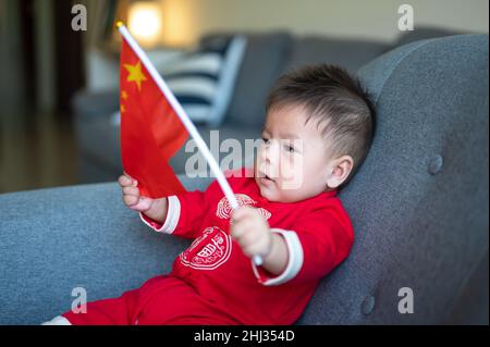 Bébé asiatique portant un drapeau chinois assis sur un canapé tout en portant des vêtements chinois traditionnels rouges pour le nouvel an lunaire à la maison Banque D'Images
