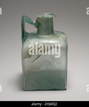 Flacon carré en verre 1st–3rd Century A.D. Roman bleu translucide, avec poignée de même couleur. Rebord replié, sur-inséré et enfoncé dans le haut de la bouche, avec surface supérieure biseautée et irrégulière ; col cylindrique, extensible vers le bas ; épaulement enfoncé avec coins arrondis et inclinés ; côtés plats et verticaux ; fond légèrement concave ;Large poignée à lanière, avec décoration peignée sur la surface extérieure, fixée au bord extérieur de l'épaule, tracée verticalement, tournée en angle aigu et traîné sur le cou, avec une traînée s'étendant sur le dessous de la jante. Décoration moulée sur le fond comprenant quatre angles en L à co Banque D'Images