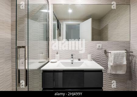 Toilette avec armoire en bois laqué noir brillant avec évier en résine blanche, miroir rectangulaire sans cadre, serviettes blanches et douche avec écran en vacances Banque D'Images