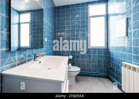 Salle de bains avec armoire en bois laqué blanc brillant avec bassin en résine blanche, miroir rectangulaire sans cadre et murs carrelés de carreaux bleu kitsch Banque D'Images