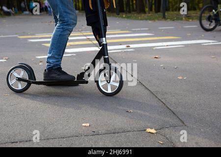 Les jambes d'un homme en jeans et baskets sur un scooter dans le parc en automne avec des feuilles jaunes sèches tombées sur l'asphalte.Promenades en automne, vie active Banque D'Images