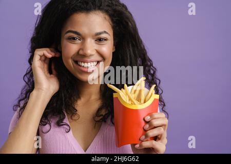 Jeune femme noire souriant tout en mangeant des frites isolées sur fond violet Banque D'Images