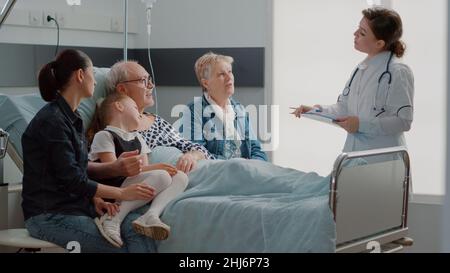Medic expliquant le diagnostic au vieux patient et à la famille en visite, assis dans le lit de la salle d'hôpital.Homme âgé parlant au médecin généraliste de traitement et de médecine, ayant des personnes en visite. Banque D'Images