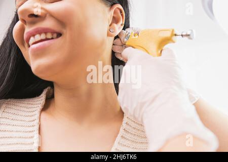 Photo rognée d'une patiente souriante qui obtient un perçage d'oreille par un spécialiste en gants médicaux dans l'armoire du cosmetologiste. Banque D'Images