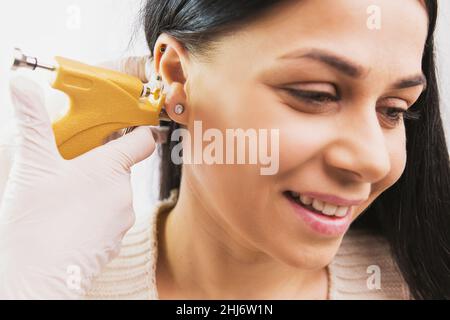 Un médecin en gants médicaux stériles perce les oreilles d'une jeune belle femme souriante dans le bureau médical à l'aide d'un pistolet à perce instantané et médical Banque D'Images