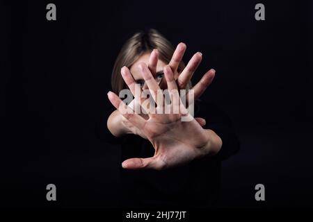 Une femme avec les mains devant elle montrant un panneau d'arrêt se cachant le visage en robe noire sur fond noir.Victime d'abus physique et psychologique Banque D'Images