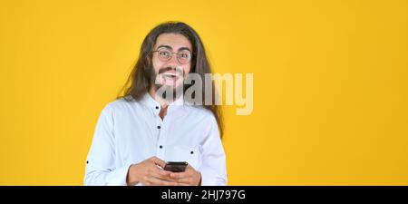 Jeune homme attrayant avec de longs cheveux dans des lunettes tient un smartphone dans ses mains et est surpris isolé sur fond jaune studio Banque D'Images