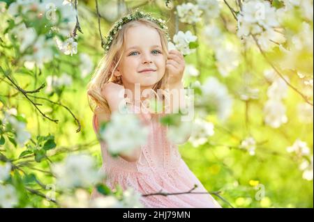 Une belle petite fille avec une couronne sur sa tête et une délicate robe rose joue dans un jardin fleuri au coucher du soleil.Un enfant heureux se reposant à l'extérieur Banque D'Images