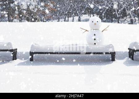 Un bonhomme de neige fait joli et mignon sur une chaise de parc dans la neige déversante est un paysage confortable et émotionnel. Banque D'Images
