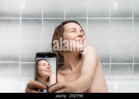Une femme d'âge moyen, de quarante ans, prend un bain et prend un selfie avec son téléphone.Une femme agréable se détendant dans la salle de bains. Banque D'Images