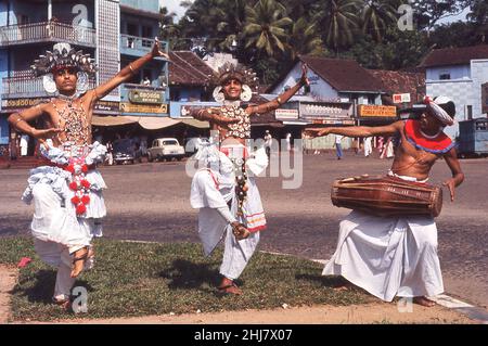 1960s, historique, Ceylan, à l'extérieur d'une place du village, sur une bordure d'herbe, deux hommes adultes en costumes, coiffures et fleurs décorées, exécutant la danse traditionnelle de Kandyan, accompagné d'un batteur jouant le gata bera, le tambour traditionnel de srilankan.La danse de Kandyan a une longue histoire dans le pays et le rituel est exécuté avec la croyance que ses bénit les gens avec la prospérité et la longue vie.Depuis ses débuts il y a plusieurs siècles, la danse est devenue un symbole de l'identité nationale sri-lankaise. Banque D'Images