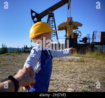 Mignon enfant dans le casque de construction tenant la main d'homme de pétrole et pointant vers le cric de pompe à pétrole.Suivez-moi la photo d'un adorable enfant qui mène le père au champ d'huile avec la machine à bascule de pompe à huile. Banque D'Images