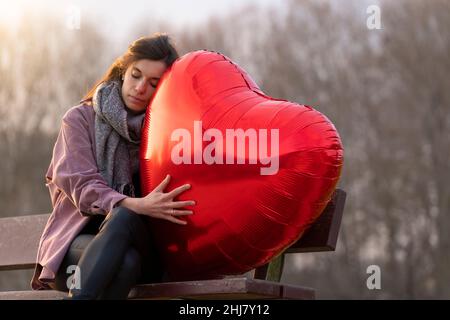 Triste jeune femme embrassant un ballon en forme de cœur assis sur un banc dans le parc Banque D'Images