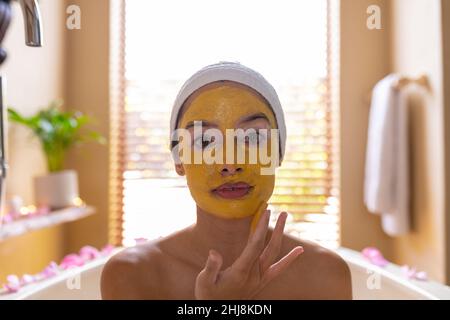 Gros plan portrait de la jeune femme biraciale appliquant un masque facial jaune dans la baignoire au spa. Non modifié, spa, soins du corps, style de vie, vitiligo et bien-être. Banque D'Images