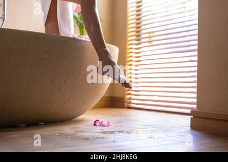 Petite section de la jeune femme biraciale avec de l'eau s'écoulant des jambes mouillées sortant de la baignoire. Non modifié, spa, soins du corps, style de vie et bien-être. Banque D'Images