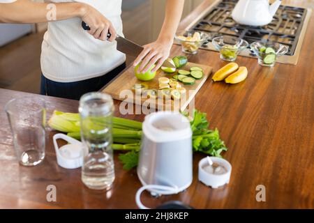 Section médiane de la jeune femme biraciale hachant des fruits tout en faisant du jus sain dans la cuisine à la maison. Non modifié, mode de vie et concept alimentaire sain. Banque D'Images